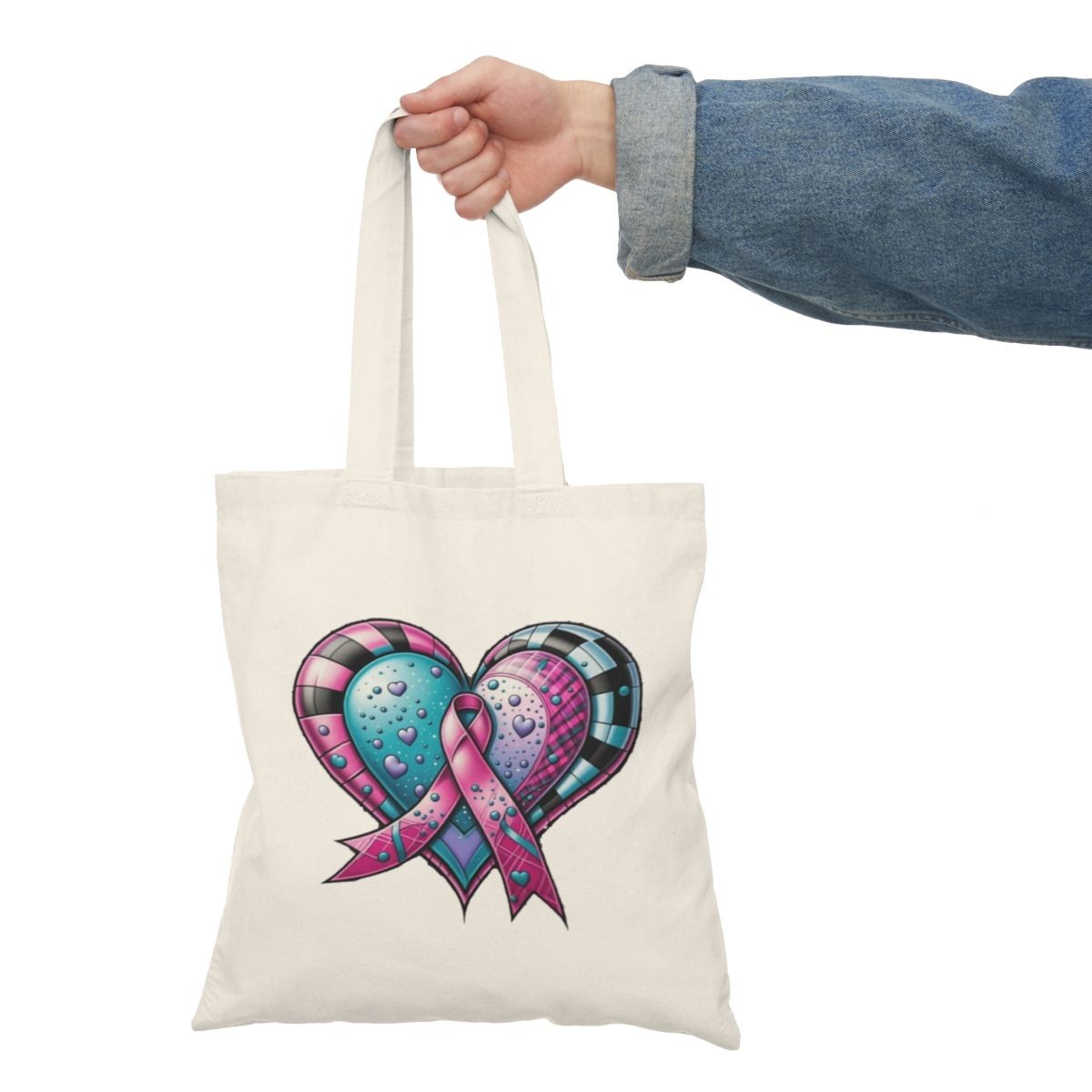 Hand holding a white tote bag with a colorful heart design and a pink ribbon in the center.